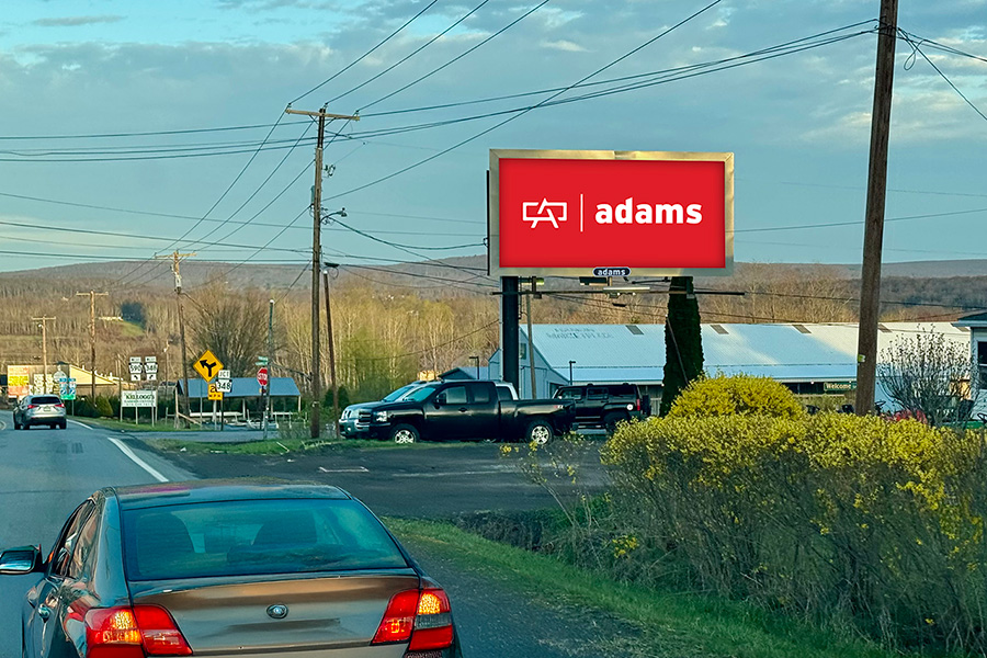 A roadside scene shows a large digital billboard with a red background displaying the text "adams" in white. Nearby, cars and trucks are parked beside a road lined with bushes. The background features hills under a partly cloudy sky, creating a tranquil rural atmosphere.