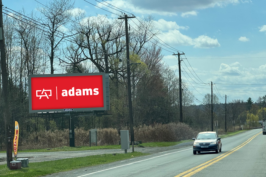 A rural road under a partly cloudy sky, with a large red billboard on the left displaying the word "adams" in white. A car drives along the road.