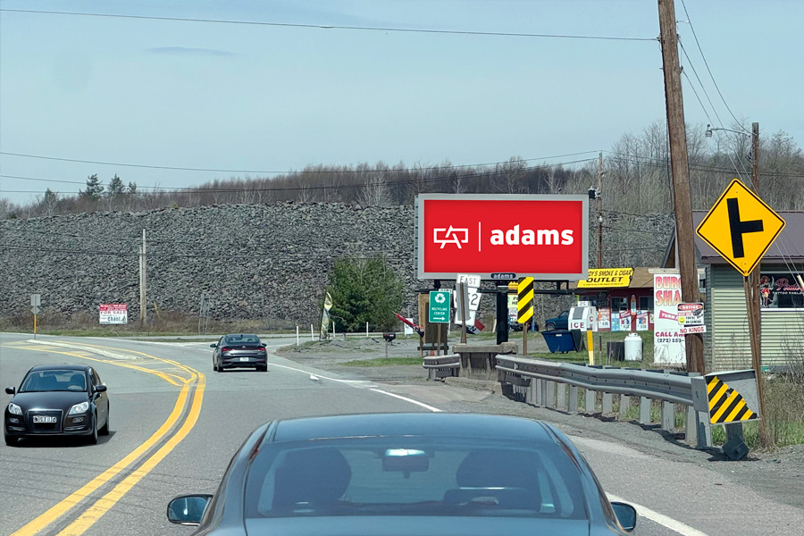 A road with three cars traveling. A red billboard with the text “adams” and a graphic is on the roadside near a yellow left turn sign.