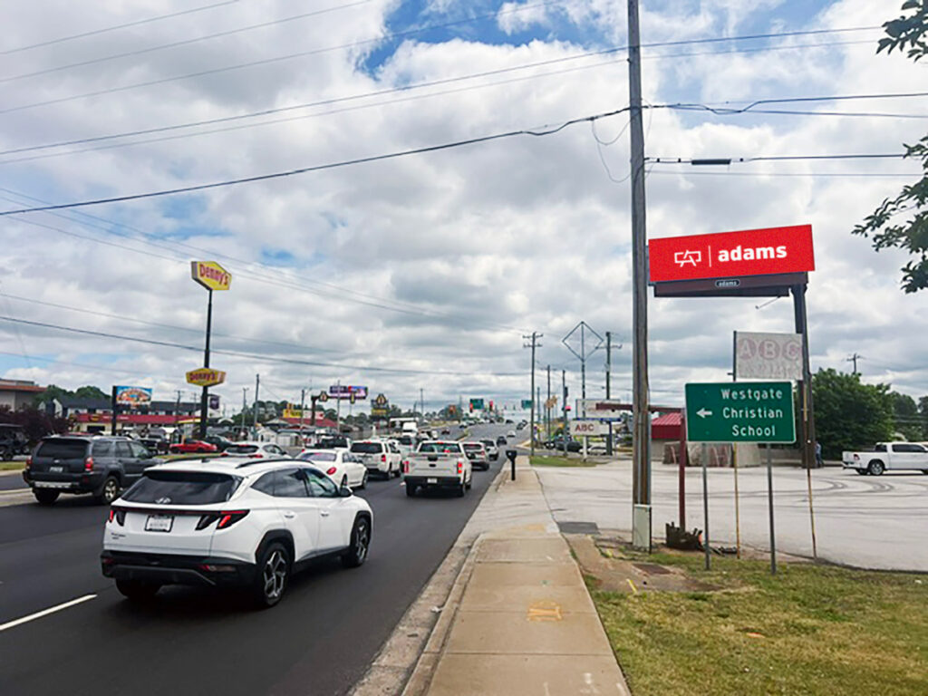 A busy road under a clear sky, with cars driving past storefronts. A red billboard reads "adams." The scene conveys a typical urban setting.