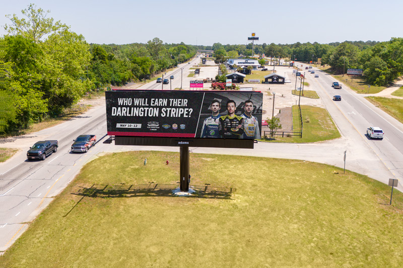 A roadside Adams Outdoor billboard with images of three race car drivers and text reading "Who Will Earn Their Darlington Stripe? May 10-12." Cars drive nearby.