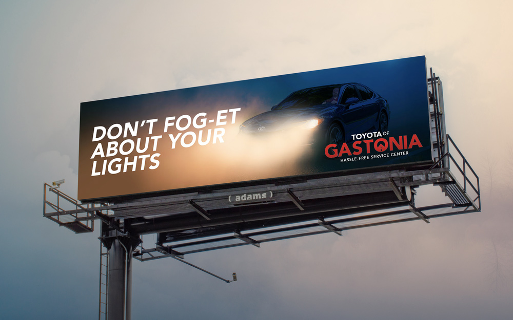 Billboard showing a car with fog lights on. Text reads, "Don't fog-et about your lights," promoting Toyota of Gastonia's hassle-free service center.