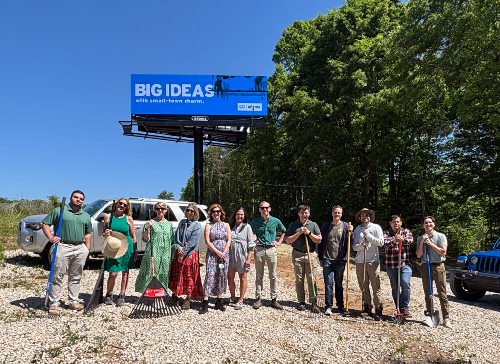 Adams Outdoor Charlotte team holding rakes and shovels below an Adams billboard after planting flowers for Earth Day