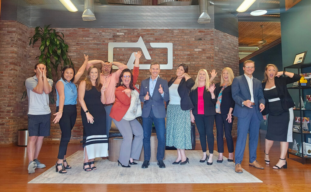 Adams Outdoor CEO Kevin Jones and the Peoria, Illinois team striking playful poses in front of the Adams logo on a brick wall