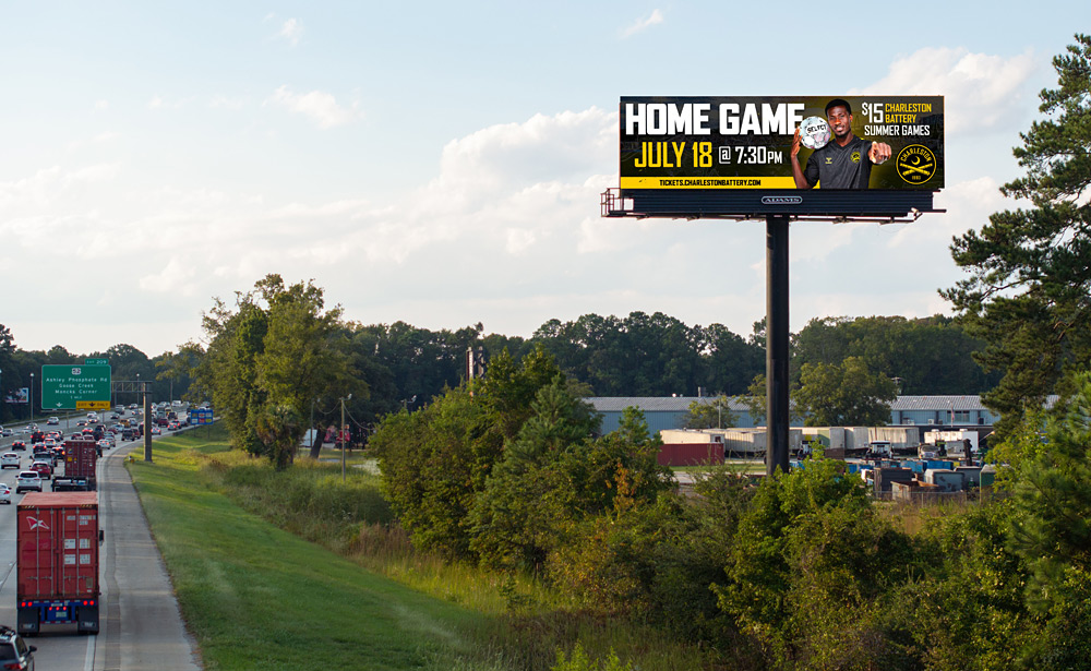 Charleston Battery 'Home Game July 18' billboard on a tall Adams Outdoor digital billboard above I-26 traffic in North Charleston.